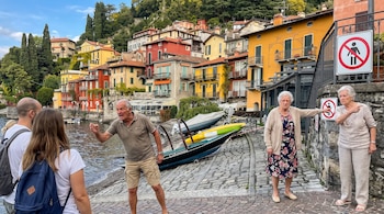 Costa de Varenna con cinco personas, edificios coloridos y botes. Tres residentes gesticulan a dos turistas cerca de carteles de prohibición de traje de baño.