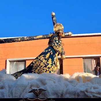 Multitud sigue un anda procesional grande con una estatua de Jesús cargando la cruz, figuras celestiales y nubes. Hay personas grabando con sus teléfonos bajo cielo azul