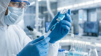 Científico con bata blanca, mascarilla y gafas protectoras sostiene un vial de líquido azul y una jeringa en un laboratorio con equipo médico al fondo.