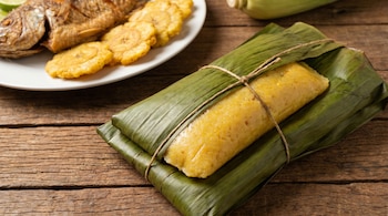 Un bollo de maíz amarillo envuelto en hojas de plátano y atado con cuerda, junto a un plato con pescado frito y patacones en una mesa de madera.