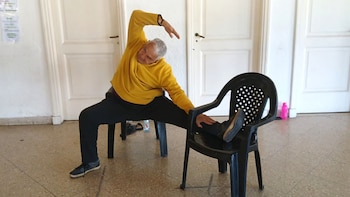 Un hombre de cabello gris con camisa amarilla y pantalones negros realiza un estiramiento lateral, apoyando una pierna en una silla negra