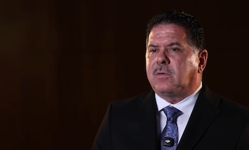 Hombre de tez blanca y bigote, con traje negro, camisa blanca y corbata azul estampada, y un micrófono clip en la corbata, con fondo oscuro