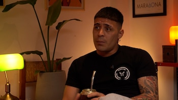 Un hombre con camiseta negra y tatuajes sostiene un mate en la mano frente a una planta de interior y lámparas de mesa