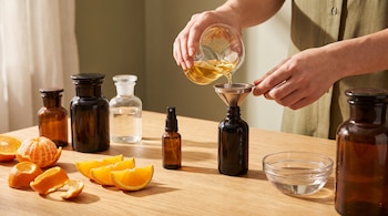 Manos vierten líquido en un frasco con embudo sobre una mesa de madera clara con cáscaras de naranja y mandarina, botellas de vidrio, un atomizador y un cuenco.