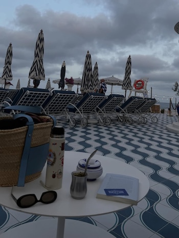 Mesa blanca con bolso, termo, gafas de sol, mate con bombilla, recipiente y libro. Fondo con sombrillas, reposeras, piso ondulado, mar y cielo nublado