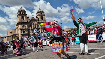 Danzarines con vestimenta tradicional de colores en desfile callejero. Una iglesia colonial de piedra y una multitud de personas en segundo plano