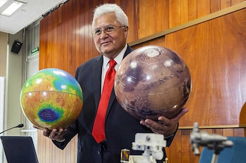 Doctor Rafael Navarro, un hombre de cabello blanco y lentes, sonríe mientras sostiene dos modelos esféricos del planeta Marte en un auditorio