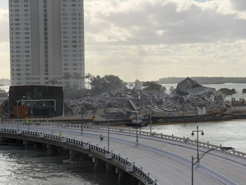 Vista aérea de los escombros de un edificio demolido en una península, junto a un puente de tráfico y un edificio alto en Miami. El cielo está nublado