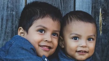 Primer plano de dos niños sonriendo frente a un fondo de madera: un niño pequeño a la izquierda y un bebé a la derecha, ambos con camisas de mezclilla