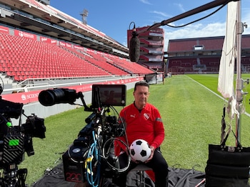 Peter Robertson, con camiseta roja de C.A.I., sentado con un balón de fútbol frente a equipo de cámara en un estadio con asientos rojos vacíos
