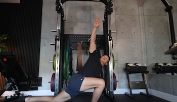 Un hombre realiza un lunge con giro de torso en un gimnasio. Se ven un rack de pesas, discos de colores, banco de ejercicio y mancuernas ajustables