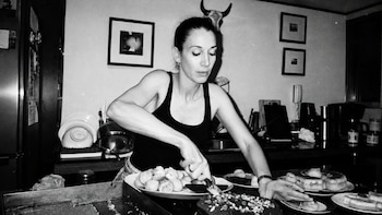 Fotografía en blanco y negro de una mujer que corta una torta sobre la mesada de una cocina con platos de comida
