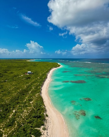 Vista aérea de una larga playa de arena blanca, aguas turquesas y arrecifes. La tierra está cubierta de vegetación verde y una casa con techo blanco se ve cerca de la playa
