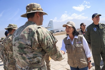 Una mujer con chaleco beige estrecha la mano de un soldado con uniforme de camuflaje en una pista de aterrizaje frente a una aeronave