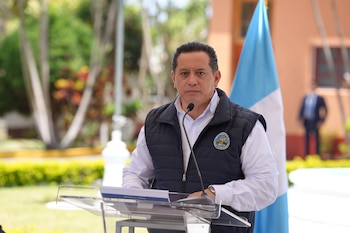 Hombre vestido con camisa blanca y chaleco azul oscuro con logo de INSIVUMEH habla en podio transparente al aire libre con bandera celeste y blanca