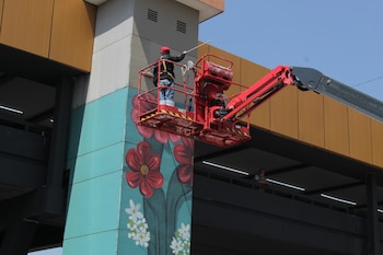 Un trabajador en un elevador de tijera rojo pinta un mural de flores rojas y blancas en una pared azul verdoso claro, con estructuras marrones y cielo azul
