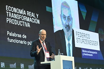 Federico Sturzenegger en traje habla en podio. Detrás, pantalla con "ECONOMÍA REAL Y TRANSFORMACIÓN PRODUCTIVA" y su retrato con nombre