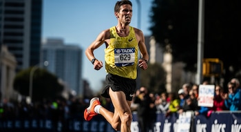 Hombre atleta con camiseta amarilla y dorsal 629, pantalones negros, zapatillas naranjas, corriendo en maratón. Expresión de esfuerzo. Fondo urbano con edificios.