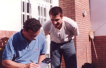 Jorge Valdano, con polo azul, sentado y firmando un libro con un bolígrafo, mientras un hombre de camiseta clara y jeans lo mira de pie. Fondo de ladrillo
