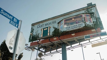 Gran valla publicitaria con diseño de casa, vegetación, cartel de calle 'Sunset Bl 8100 W'. Muestra el logo de Netflix, texto 'The Last House' y una persona en una ventana