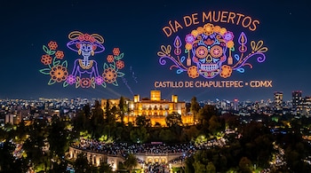 Castillo iluminado de noche rodeado de árboles y personas, bajo figuras luminosas de una calavera y una catrina formadas por drones en el cielo.