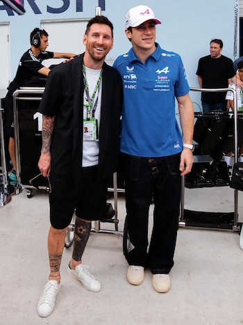 Lionel Messi y Esteban Ocon sonríen a la cámara, de pie uno al lado del otro, en un área interior con una pared clara y equipos de F1 al fondo