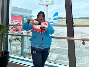 Una mujer vierte agua de un termo en un mate, con una ventana grande de aeropuerto y la cola de un avión azul y blanco al fondo