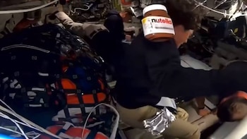 Un astronauta con camisa oscura y un tarro de Nutella flotando cerca de su hombro en el interior abarrotado de una nave espacial