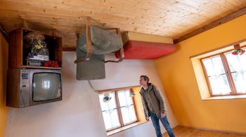 Un hombre mira hacia arriba en una habitación donde el mobiliario, incluyendo un sillón, un sofá, una televisión y un estéreo, está fijo al techo de madera. Las paredes son blancas y amarillas, con ventanas.