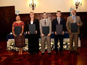 Cinco funcionarios sentados ante una mesa azul firman documentos frente a dos banderas de Guatemala y arreglos de flores blancas