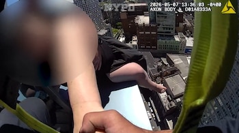 Vista desde arriba mostrando las piernas de una persona colgando del borde de un rascacielos, con edificios de la ciudad debajo. Una mano sujeta y una correa verde es visible