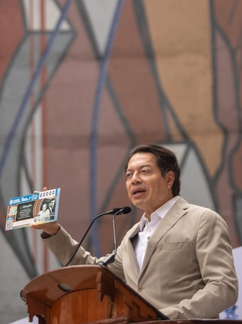 Hombre de traje claro en un podio con micrófonos, sosteniendo un billete de lotería azul. Detrás, un mural abstracto de tonos ocres y azules