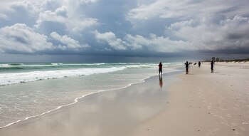 Una playa de arena blanca en Florida con cielo nublado y olas suaves rompiendo en la orilla. Pocos bañistas están de pie en la arena observando el mar.