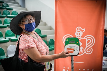 Mujer con sombrero oscuro, gafas y mascarilla azul toca un lector de tarjetas verde con una tarjeta naranja YO Jalisco, frente a un banner naranja