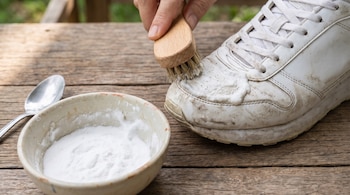 Una mano pasa un cepillo con pasta blanca sobre una zapatilla deportiva sucia, junto a un cuenco con polvo blanco y una cuchara en una mesa.