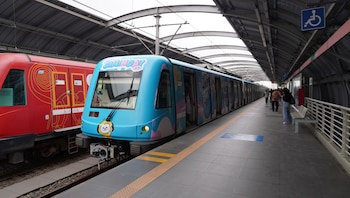 Tren azul con la inscripción Happy Jiminday y la ilustración de un personaje animado en la estación del tren junto a pasajeros en el andén