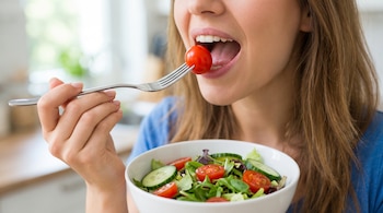 Una mujer con cabello largo y camiseta azul come un tomate cherry de una ensalada con lechuga, pepino y tomate.