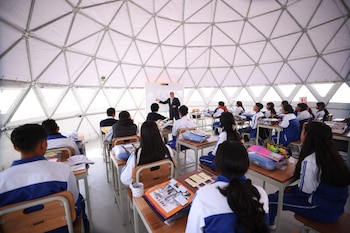 Un hombre de traje señala una pizarra frente a estudiantes sentados en pupitres dentro de un aula en forma de domo geodésico con paredes blancas