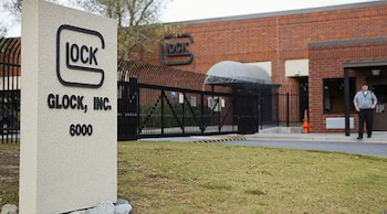 Vista exterior de la sede de GLOCK, Inc. con un cartel de piedra que muestra el logo y nombre, una valla negra, un edificio de ladrillo y un guardia