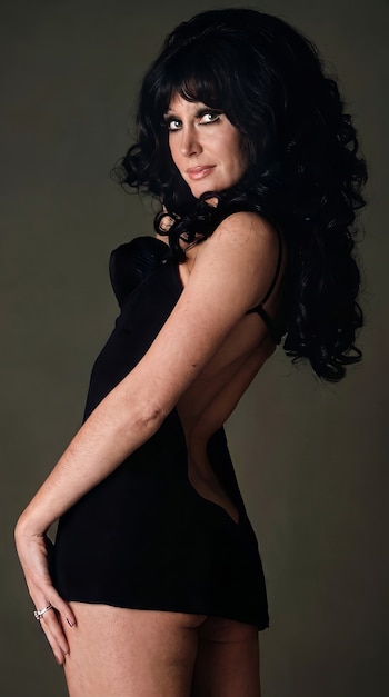Una mujer con cabello largo y oscuro y un vestido negro sin mangas posa sobre un fondo gris liso. Su brazo derecho está estirado y su espalda descubierta.