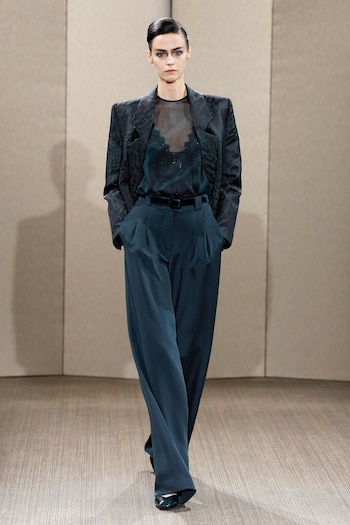 Modelo con chaqueta estampada, blusa transparente, pantalón ancho, cinturón de terciopelo y zapatos. Cabello recogido y maquillaje en los ojos. Fondo liso