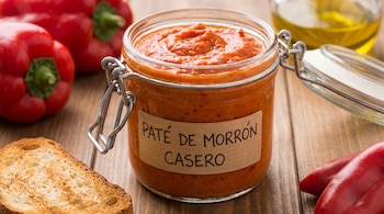 Frasco de vidrio con paté rojo etiquetado, junto a una tostada de pan, pimientos morrones y aceite de oliva sobre una superficie de madera.
