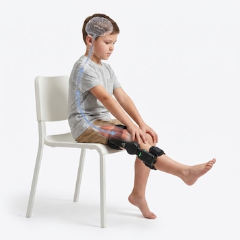 Niño de unos 7 años sentado en una silla, con un robot ortopédico negro en su pierna derecha que extiende activamente. Una flecha azul ilustra la activación cerebral.