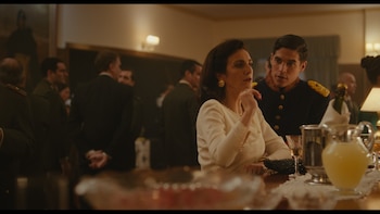 Primer plano de una mujer en crema y un hombre en uniforme militar azul con insignias doradas, conversando en un evento social, con jarra de Mimosa