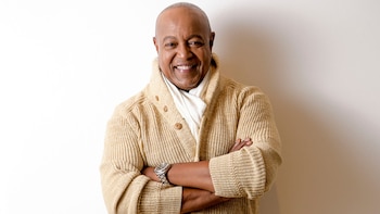 Retrato de Peabo Bryson, un hombre de piel oscura con cabeza rapada, sonriendo. Viste un cárdigan beige de punto, camisa blanca y reloj plateado. Brazos cruzados