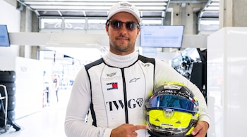 Hombre con gorra y gafas de sol, vestido con uniforme blanco y negro de piloto, sostiene un casco de carreras amarillo, negro y azul, y lo señala.