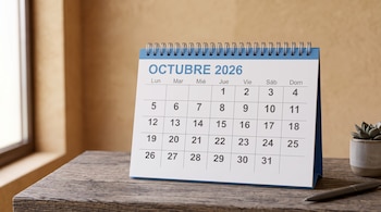 Calendario de mesa en color azul y blanco mostrando octubre de 2026 sobre una mesa de madera con un bolígrafo y una maceta.