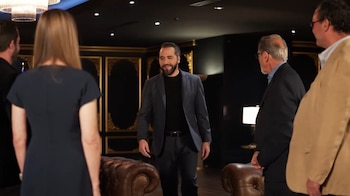 Un hombre barbudo en traje oscuro gesticula sentado en un sillón de cuero, rodeado de tres personas en un lujoso salón con banderas de El Salvador y un candelabro