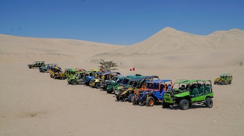 Una hilera de vehículos tubulares de distintos colores sobre la arena con dunas al fondo y cielo despejado.