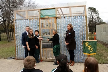 Cinco personas, incluyendo dos niños con uniforme escolar, cortan una cinta inaugural frente a una estructura hecha de botellas de plástico
